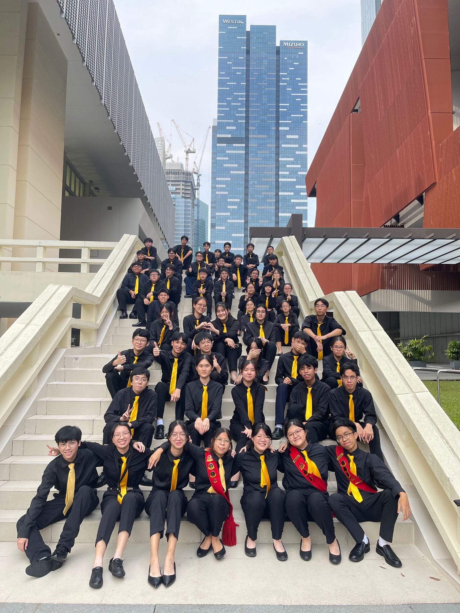 NV Symphonic Band members in black uniforms with yellow ties posing on outdoor stairs with tall city buildings behind them.